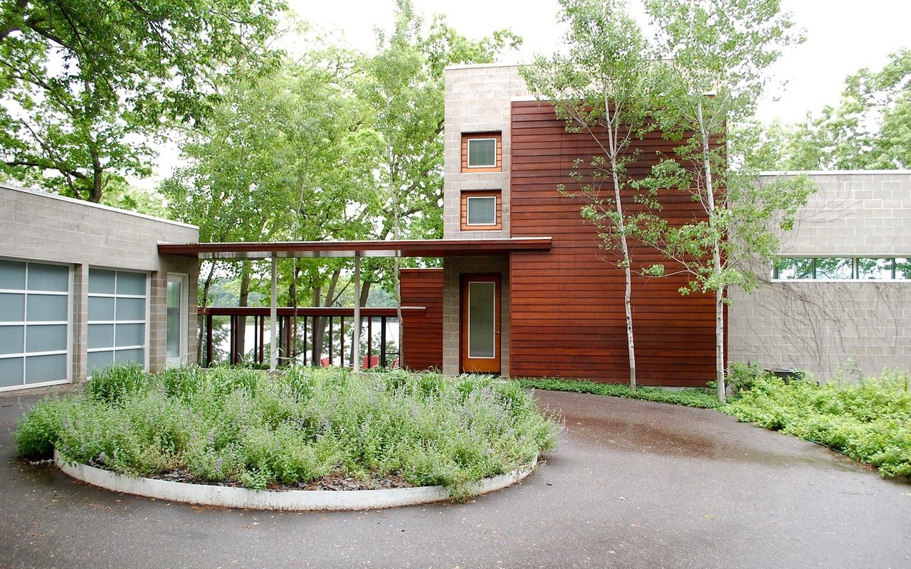 Modern home with red wood siding and a covered walkway, framed by leafy trees and landscaped gardens.