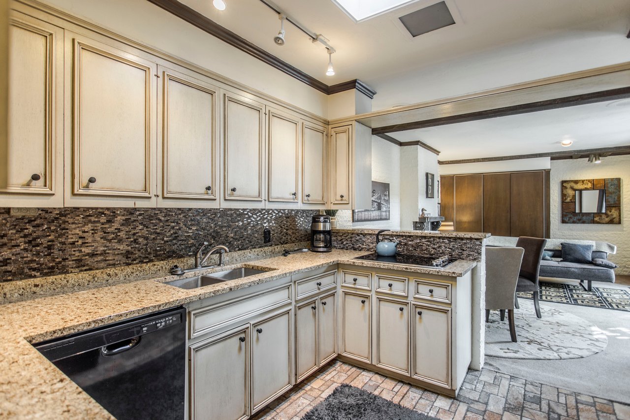 Kitchen with cream paneled cabinets, granite counters, and a dark mosaic backsplash opening to a living area.