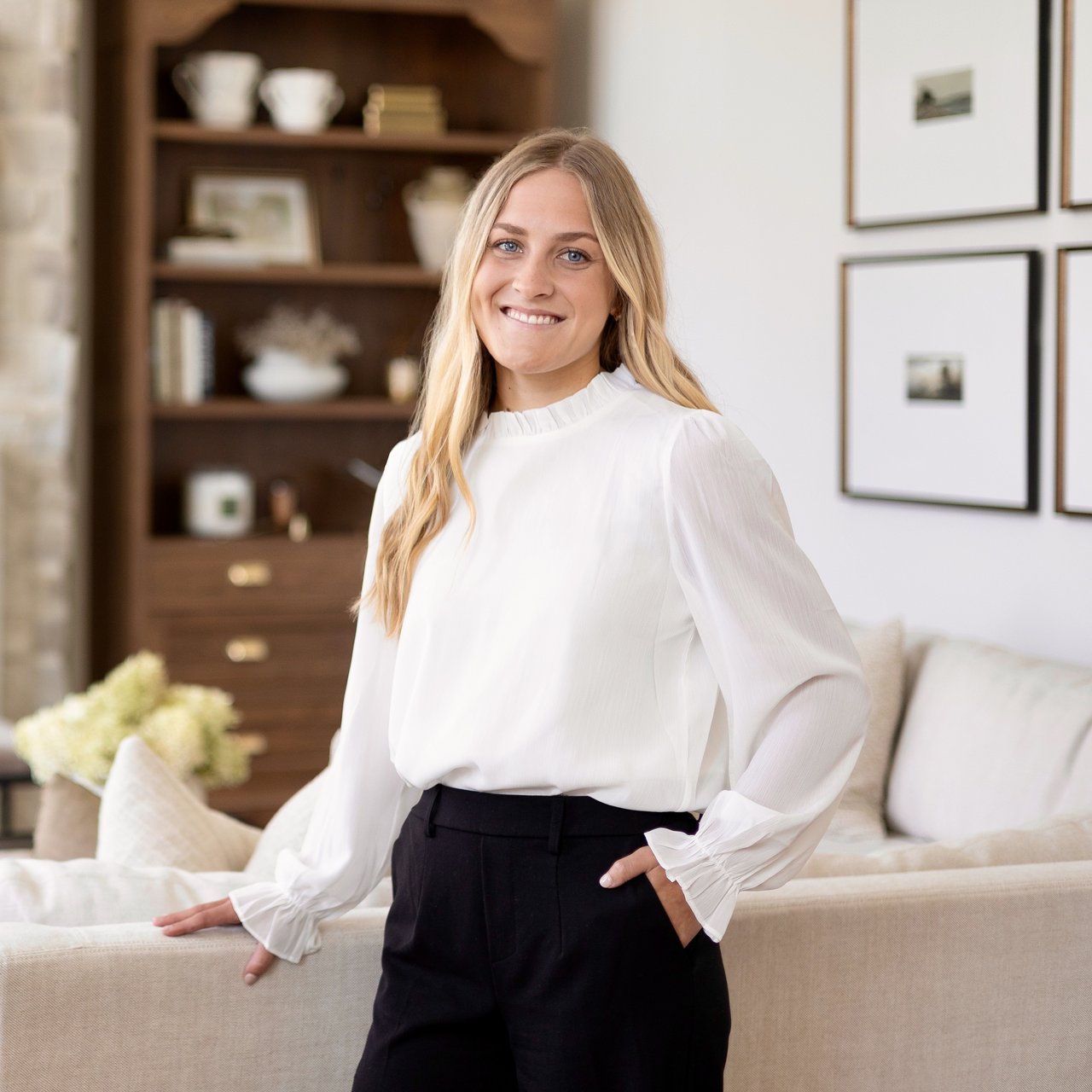 Anna Markham in a white blouse and black trousers beside a living room sofa.