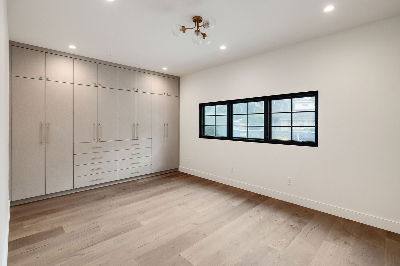 Empty bedroom with built-in wardrobes, light wood flooring, and a wide black-framed window.