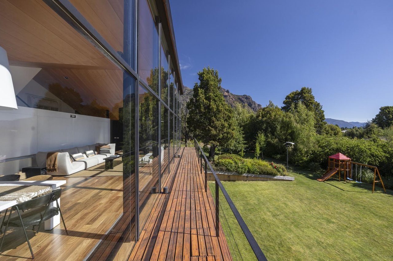 Modern glass-fronted home with a timber deck overlooking a garden, playset, and mountains.
