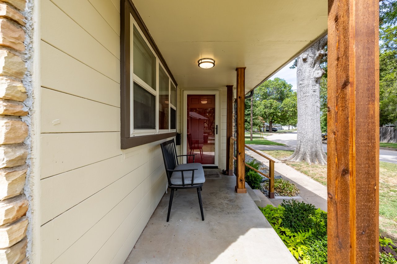 Covered front porch with cream siding, wooden posts, and a black chair overlooking a leafy street.