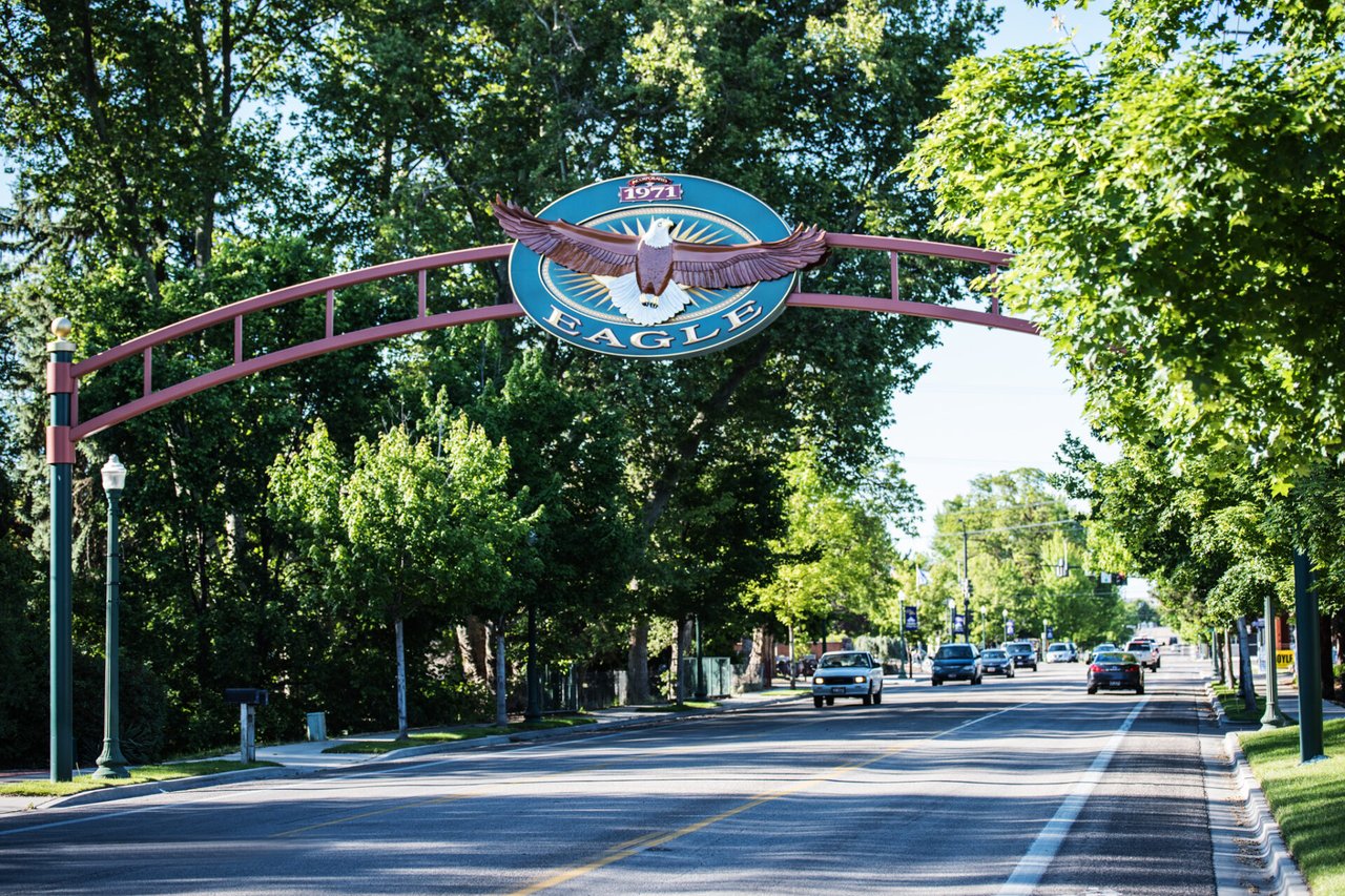 Eagle entrance sign arches over a sunny, tree-lined street with passing cars.