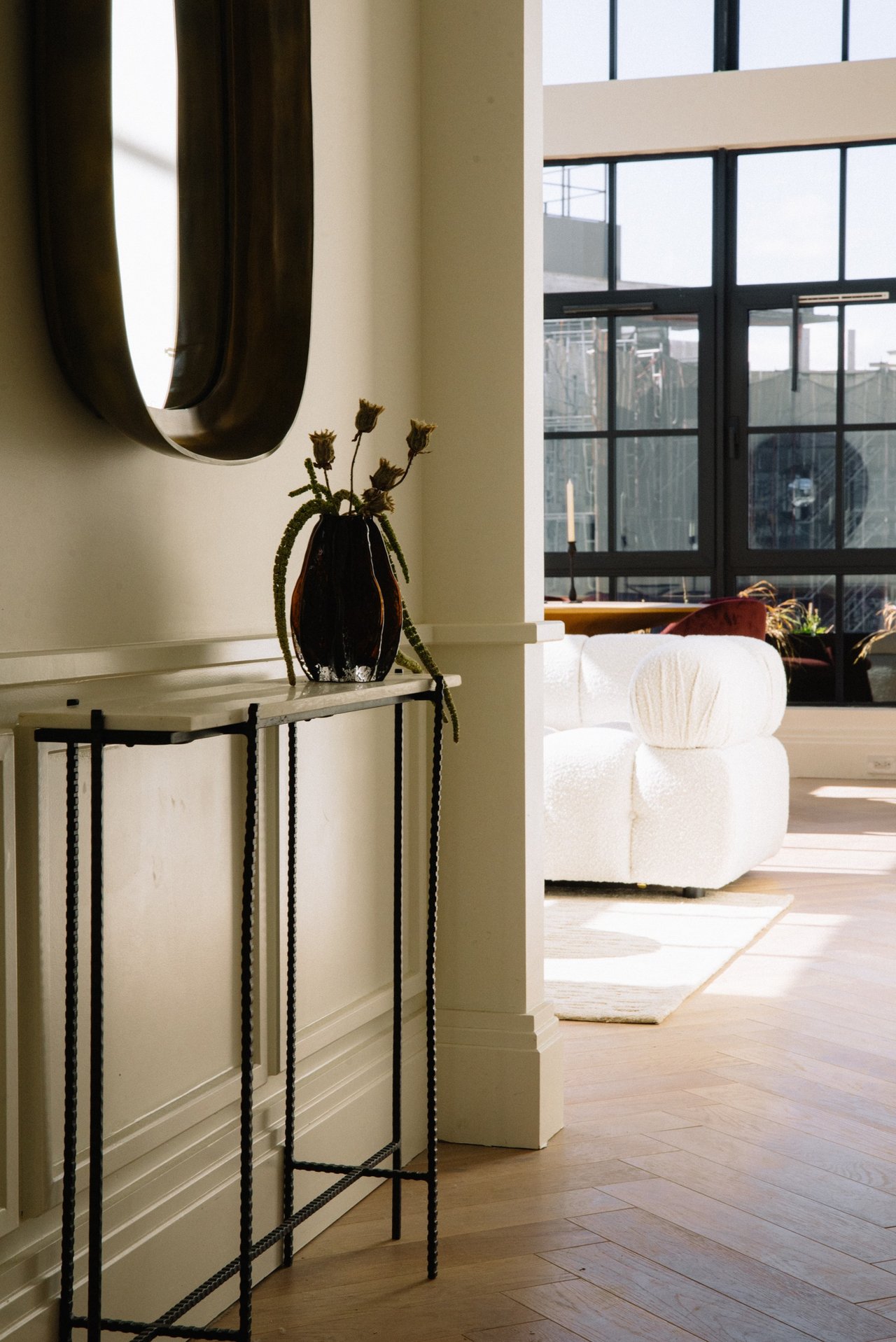 Staged apartment living room with a white sculptural chair, tall black-framed windows, and herringbone floors.