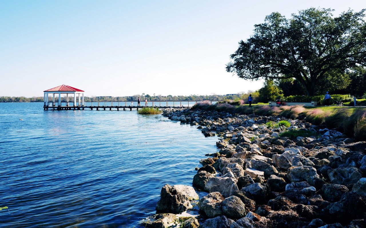 Waterfront park with a red-roofed gazebo on a pier and a rocky shore beneath a large oak tree.
