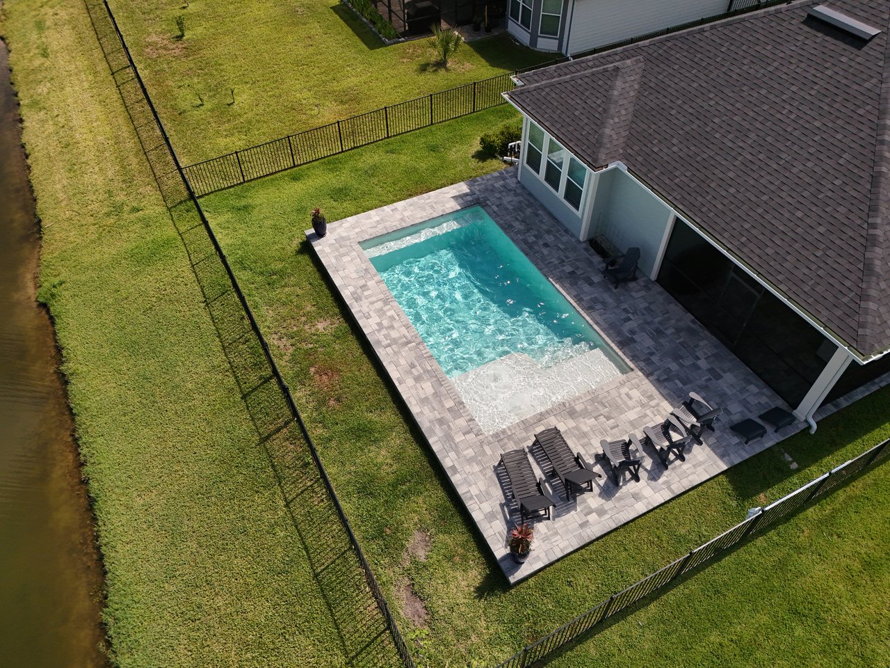 Aerial view of a home with a rectangular pool, paved patio, fenced lawn, and canal.
