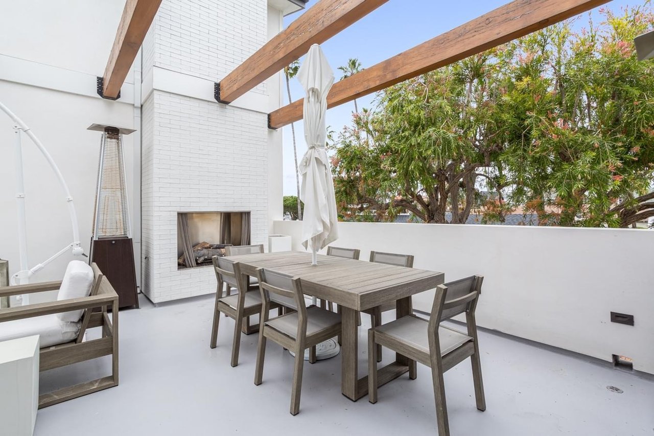  A contemporary outdoor patio with a dining table, a white brick fireplace, a space heater, and wood beams overhead.