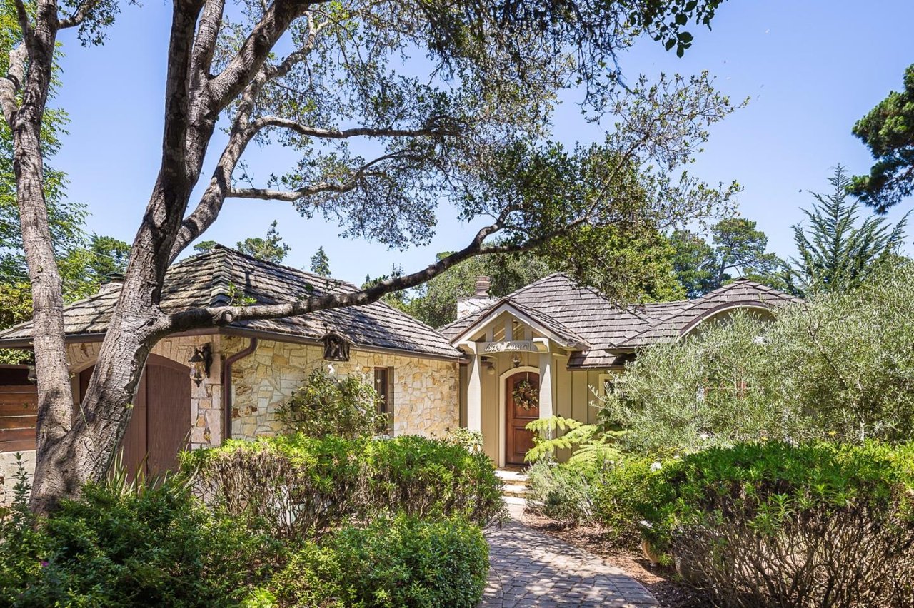 Stone-and-wood home framed by mature trees, with landscaped shrubs and a curved walkway to the entrance.