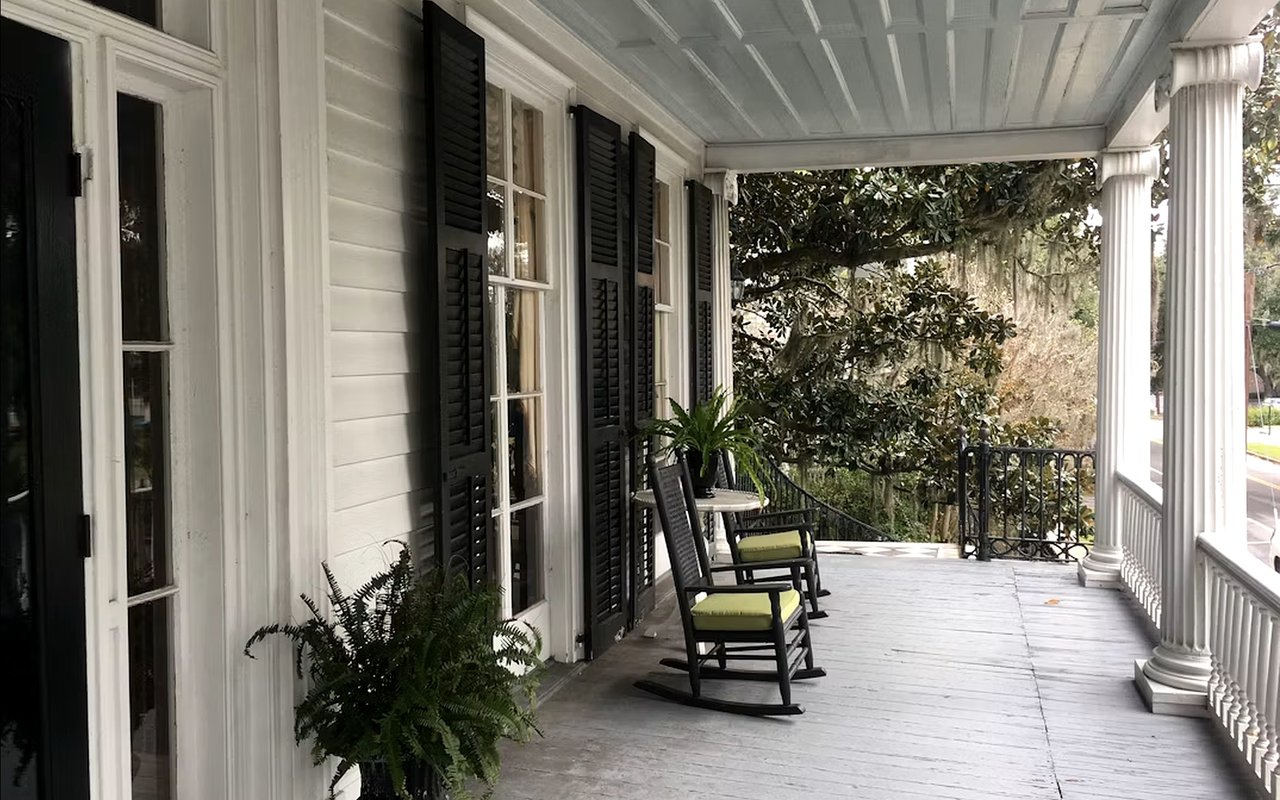 Covered front porch with white columns, black shutters, potted ferns, and green-cushioned rocking chairs.