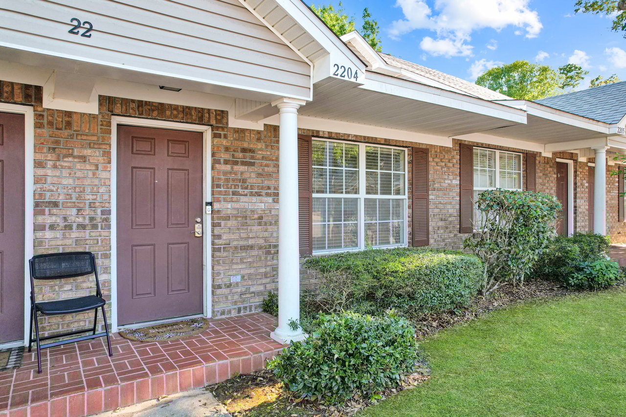Front view of a brick house with a brown door, covered porch, white columns, and windows with shutters. A black chair sits on the red-tiled entryway. Well-kept shrubs and lawn contribute to a welcoming atmosphere under a clear sky.