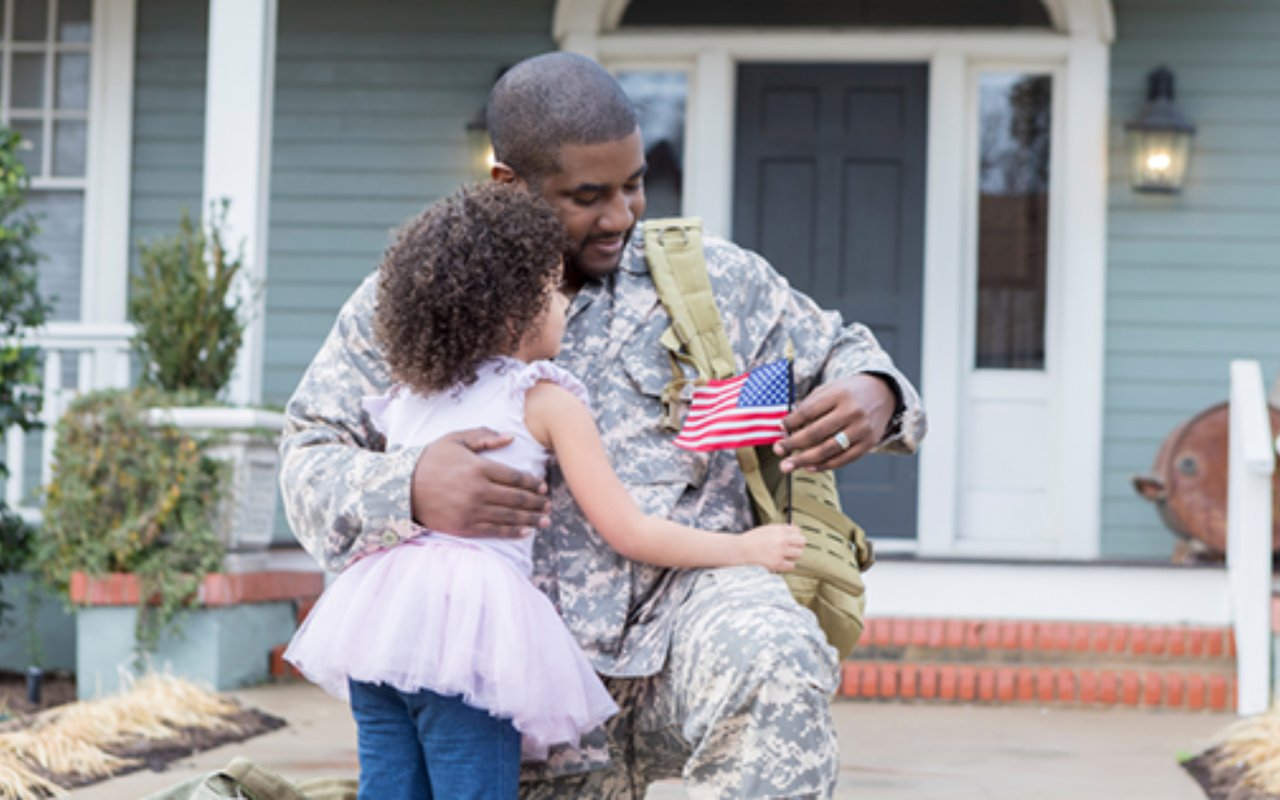A servicemember embraces a young girl holding an American flag outside a blue house.