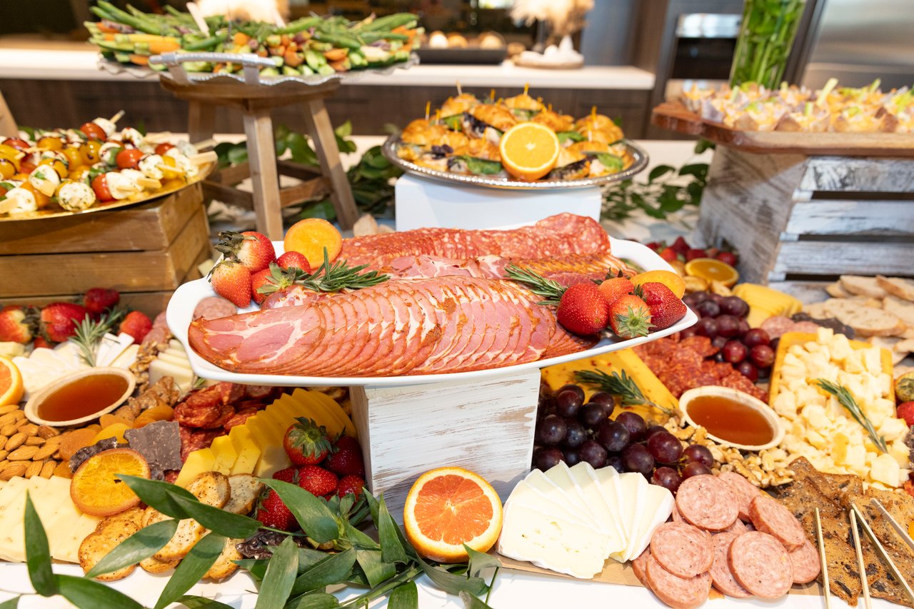 Buffet spread with sliced ham, cheeses, fruit, crackers, and assorted appetizers.