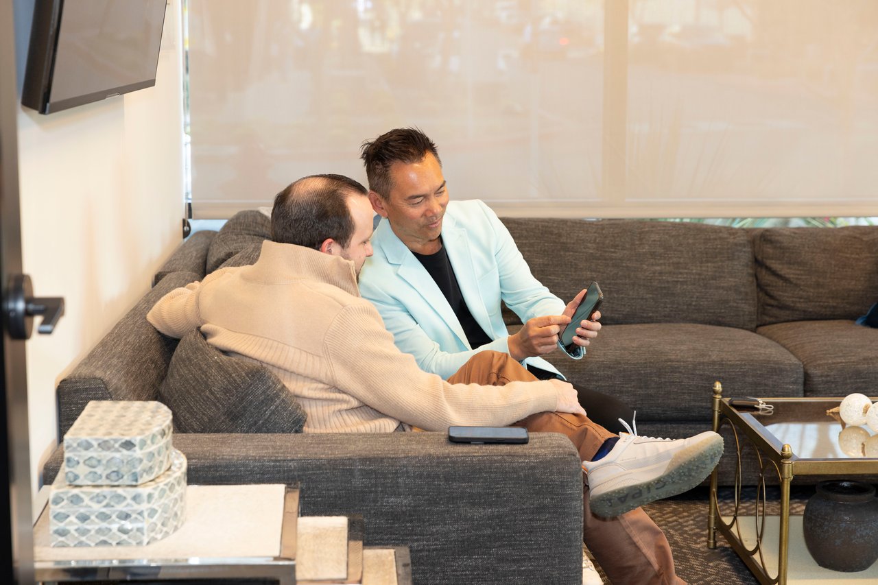 Two men sit on a sectional sofa and look at a smartphone; one wears a light blue blazer.