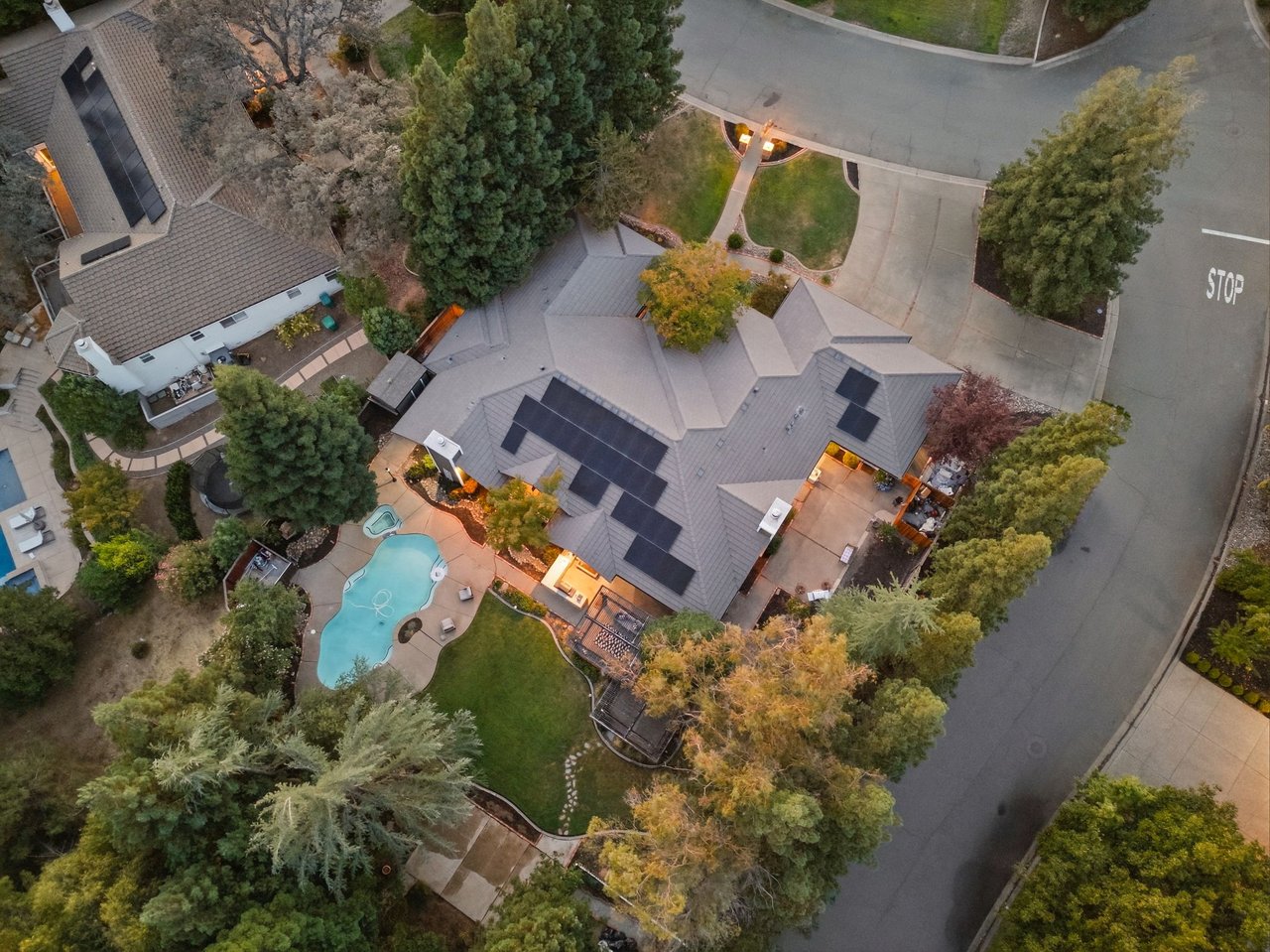 Aerial view of a home with rooftop solar panels, a curved pool, and mature trees.