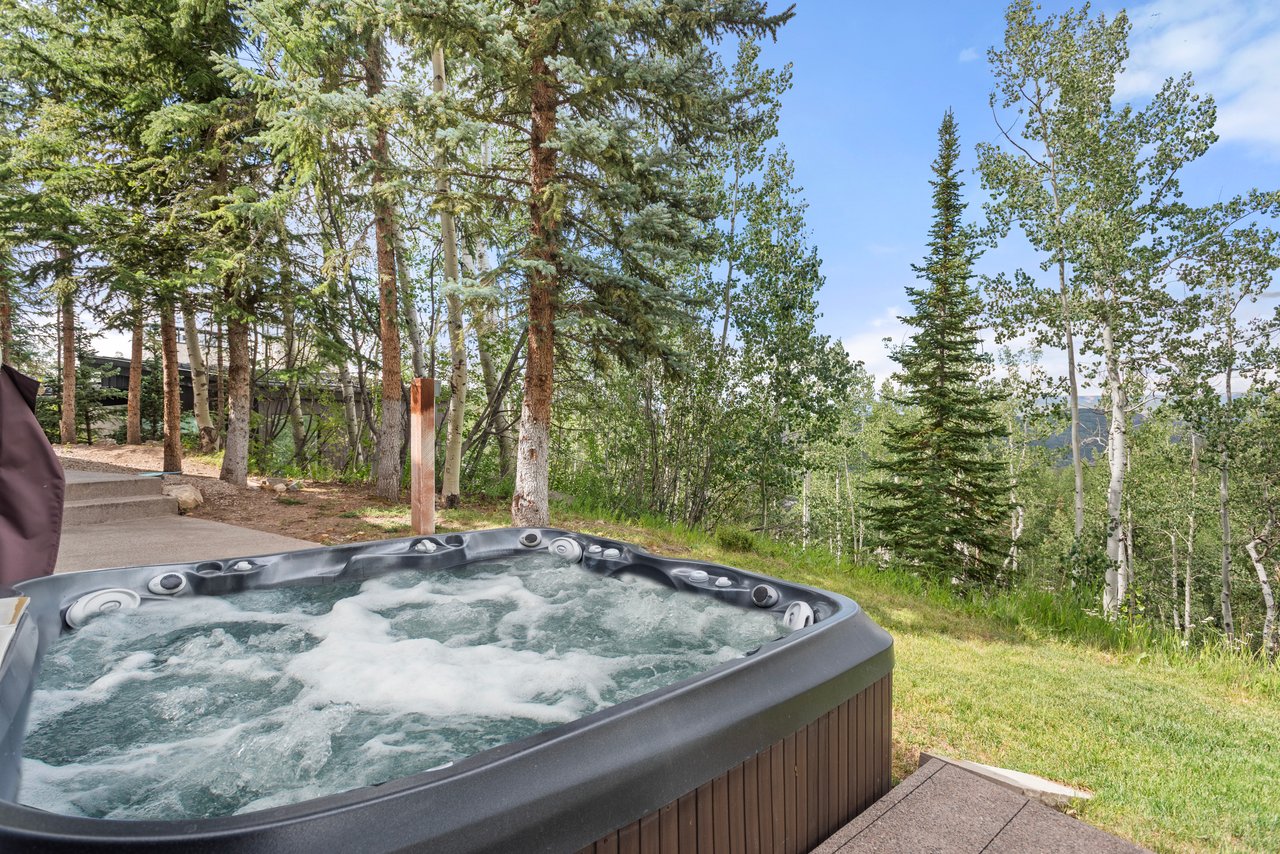 Outdoor hot tub beside a wooded patio, with evergreens, aspens, and a distant mountain slope.