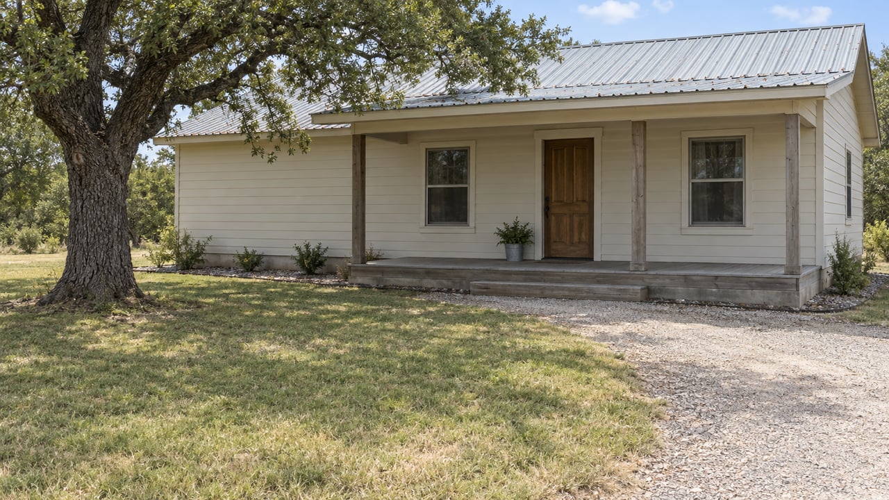 Modest wood-sided home with a galvanized metal roof, gravel approach and mature post oak beside a gently sloping grass yard.