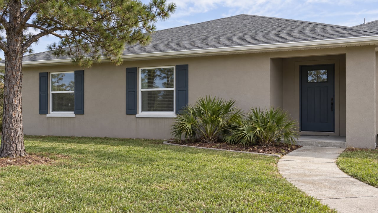 Taupe single-story home with a gray hip roof, blue shutters, curved concrete walkway and broad green front lawn.