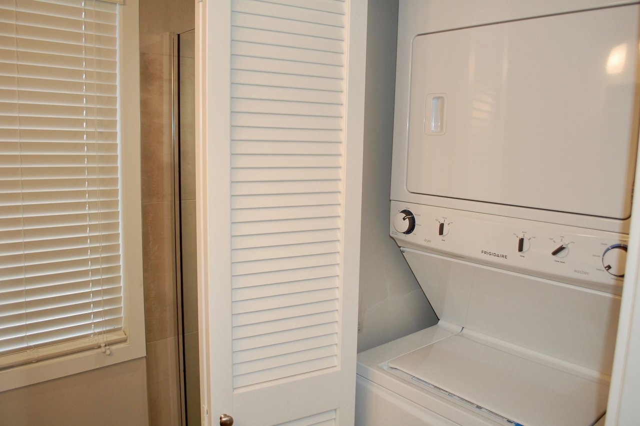 Stacked white washer and dryer beside a louvered closet door and window blinds.