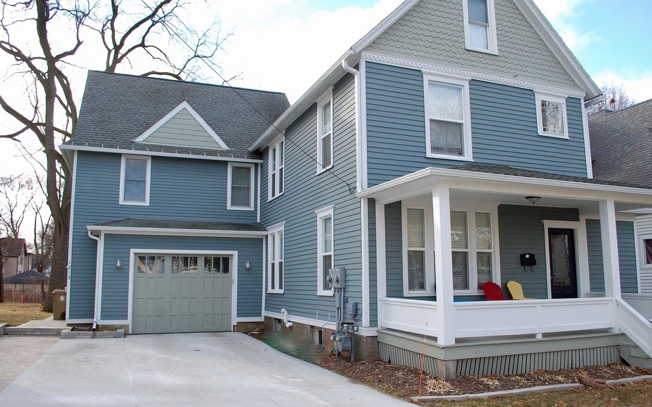 Blue two-story house with white-trimmed porch, red and yellow chairs, and attached garage.