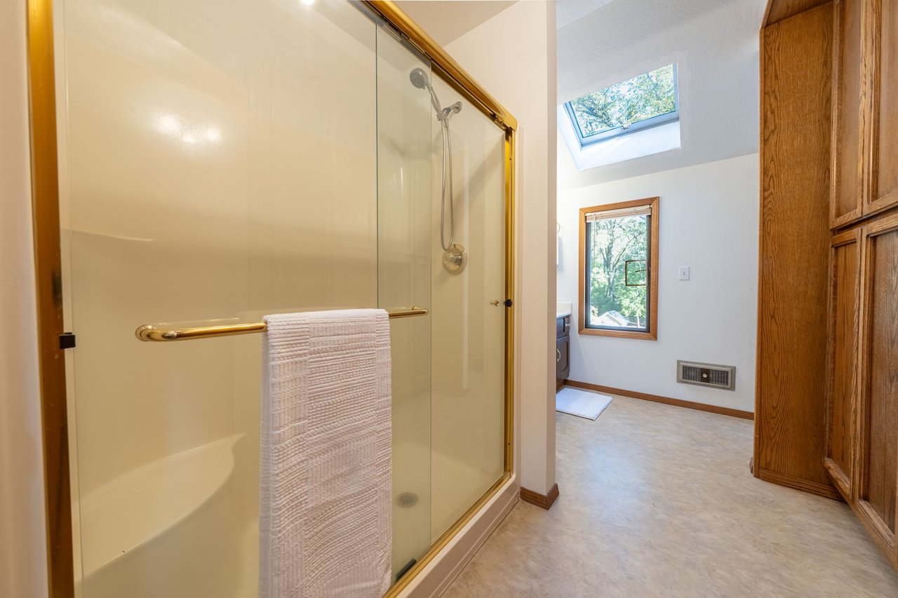 Bathroom with a gold-trimmed glass shower, skylight, wood-framed window, and built-in cabinetry.