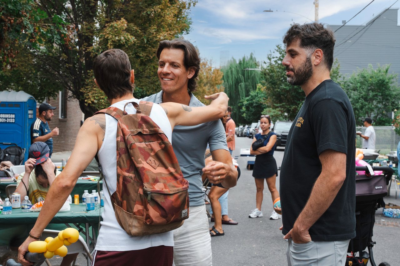 Neighbors greet one another at a residential street block party with tables and food nearby.