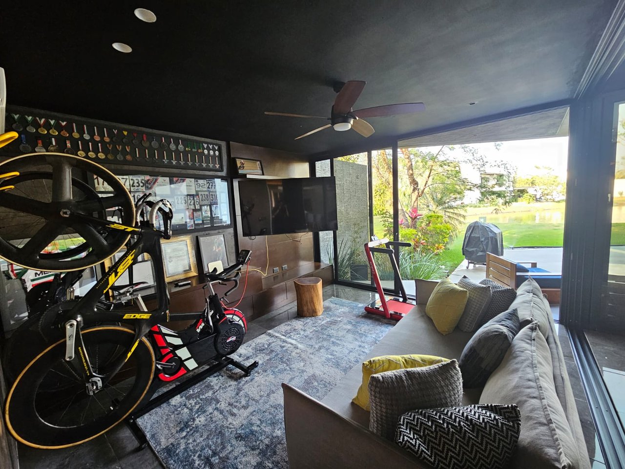 Recreation room with cycling equipment, sofa seating, and glass doors overlooking a lawn and pond.