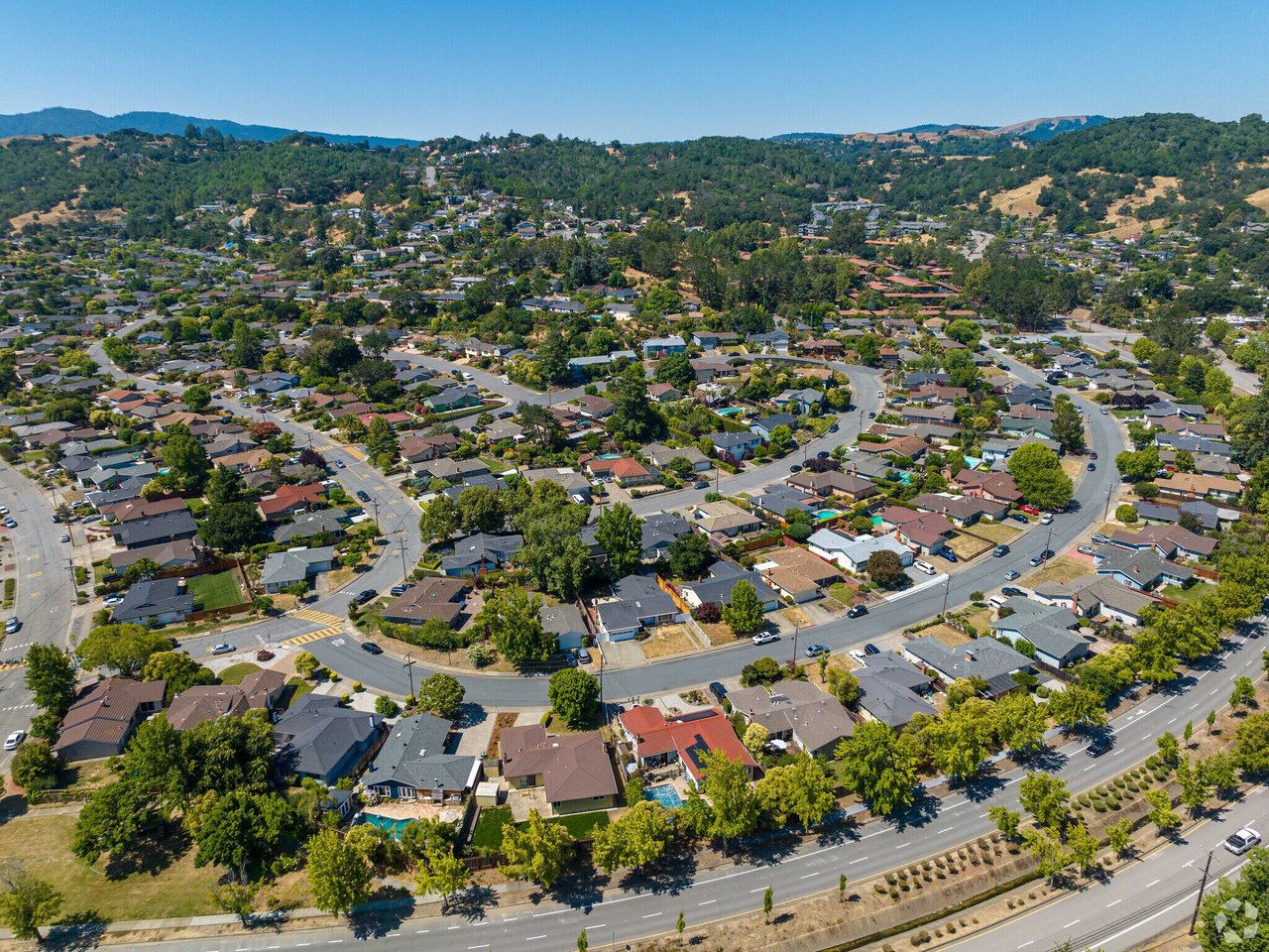 Aerial view of a tree-lined neighborhood of detached homes, with wooded hills beyond.