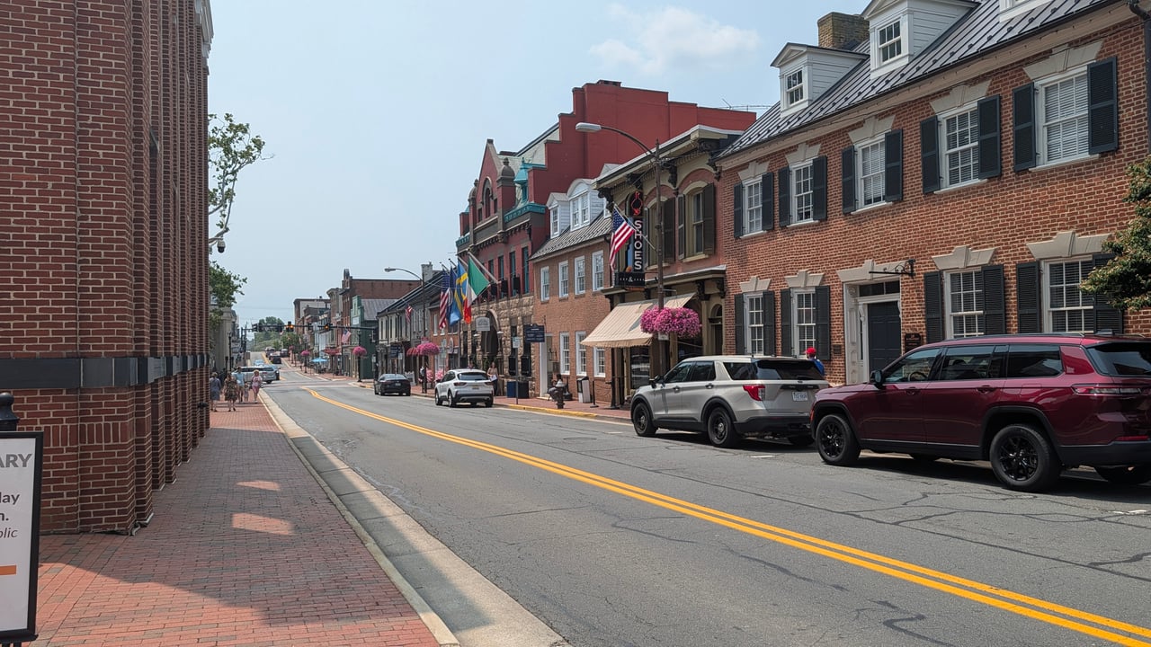 A street-level perspective looking down the paved corridor past historic brick storefronts, colorful awnings, hanging flower baskets, and parked cars in downtown Berryville, VA, documented by local real estate expert Diana Geremia.