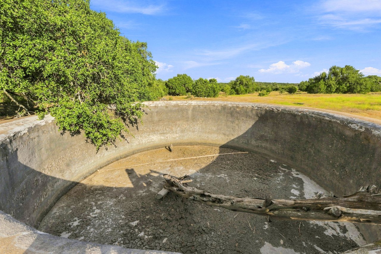 Empty circular concrete basin with fallen branches, beside trees and open ranch grassland.