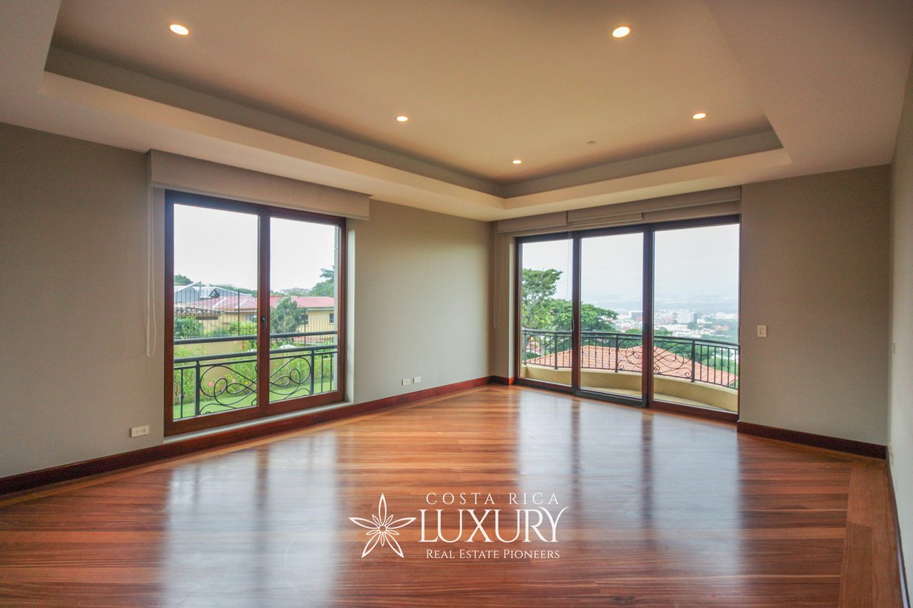 Vacant apartment living area with polished wood floors, large windows, and a balcony overlooking greenery and the city.