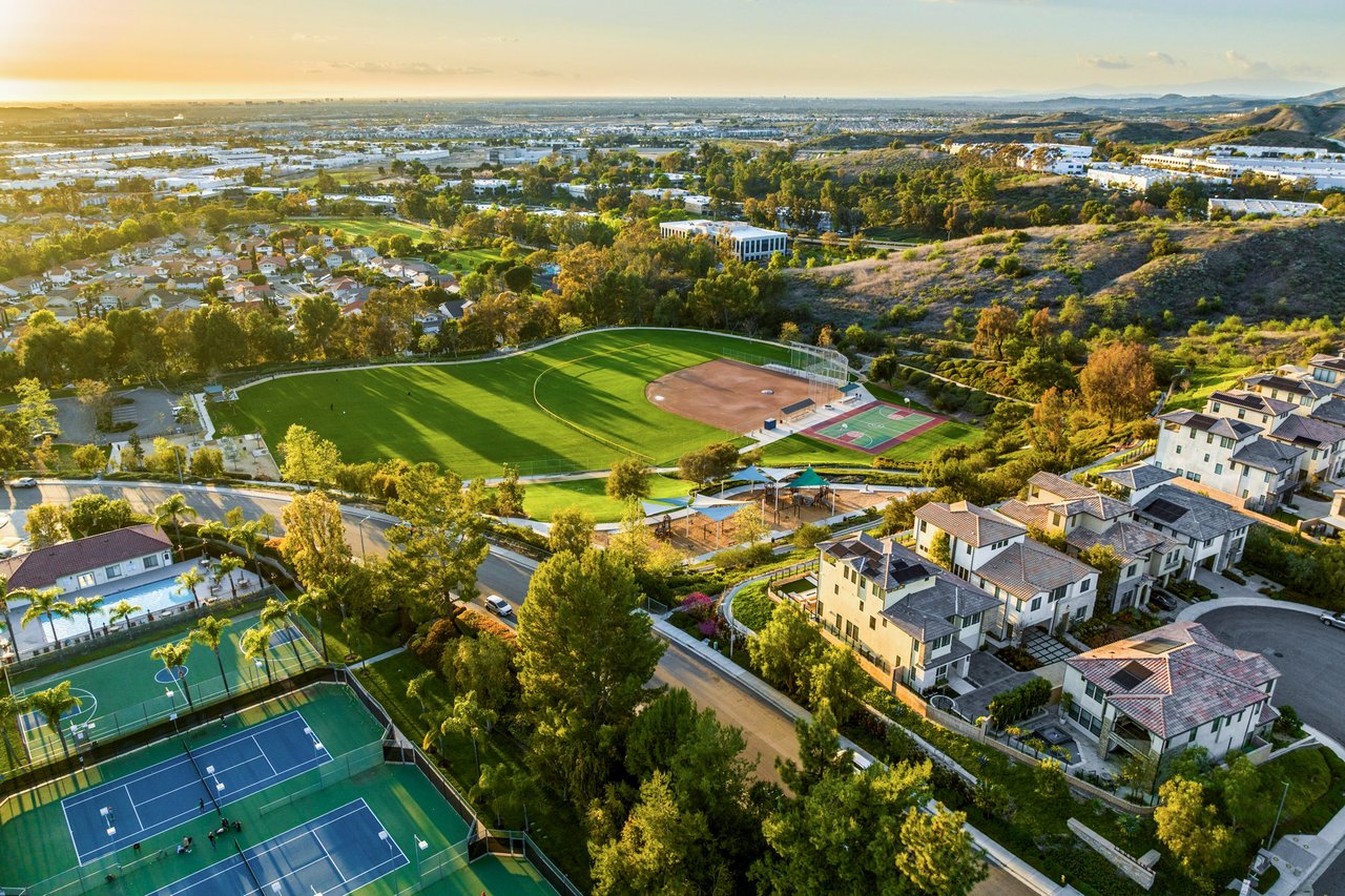 Aerial view of homes surrounding tennis courts, a baseball field, and green parkland.