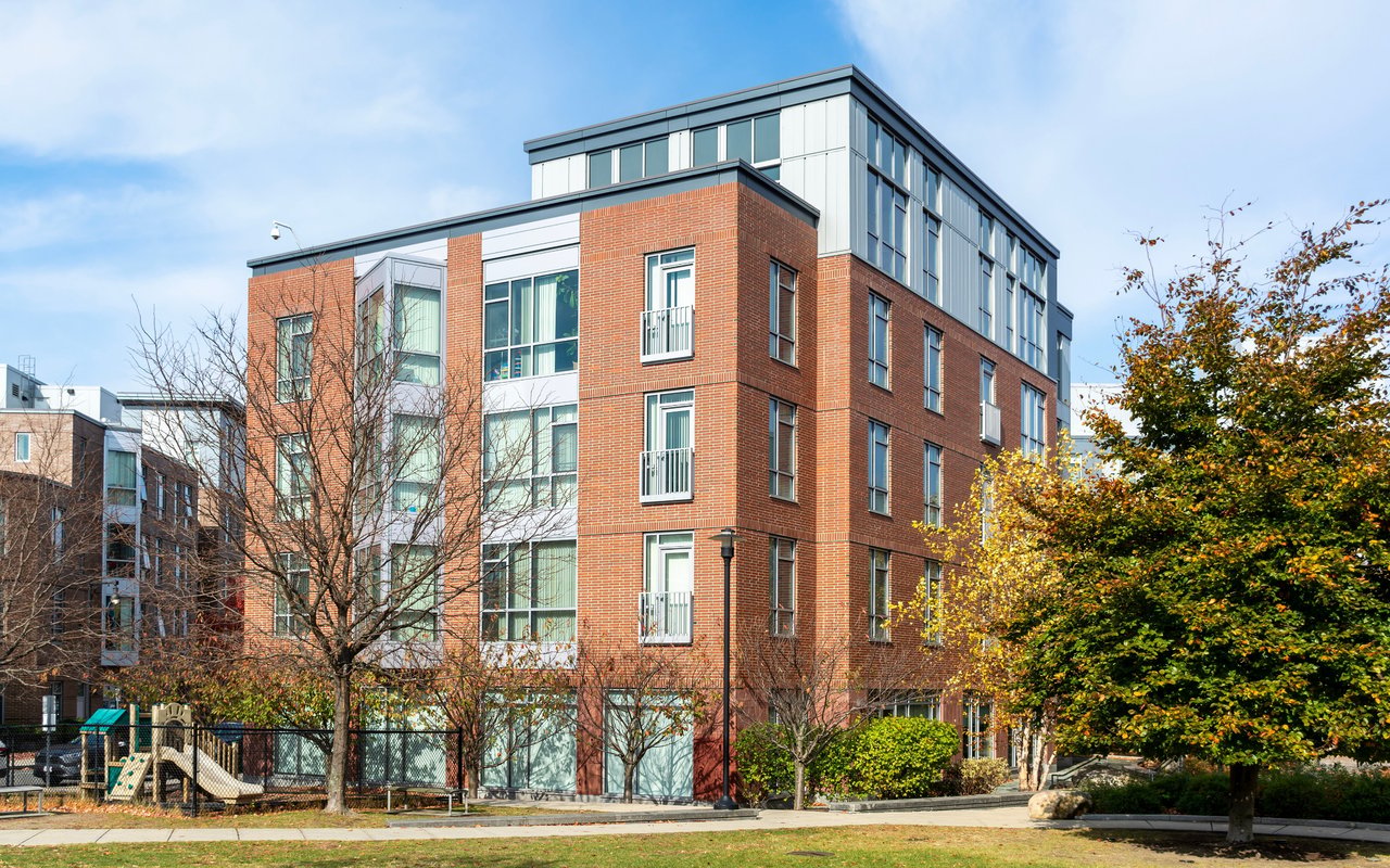 Modern brick-and-glass apartment building with balconies, landscaped grounds, and a playground.