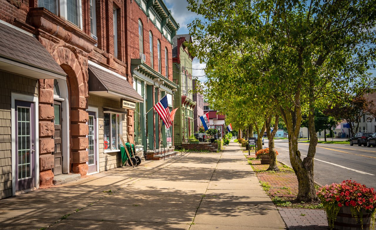 Downtown Perry storefronts along a tree-lined sidewalk, with historic brick façades and American flags.