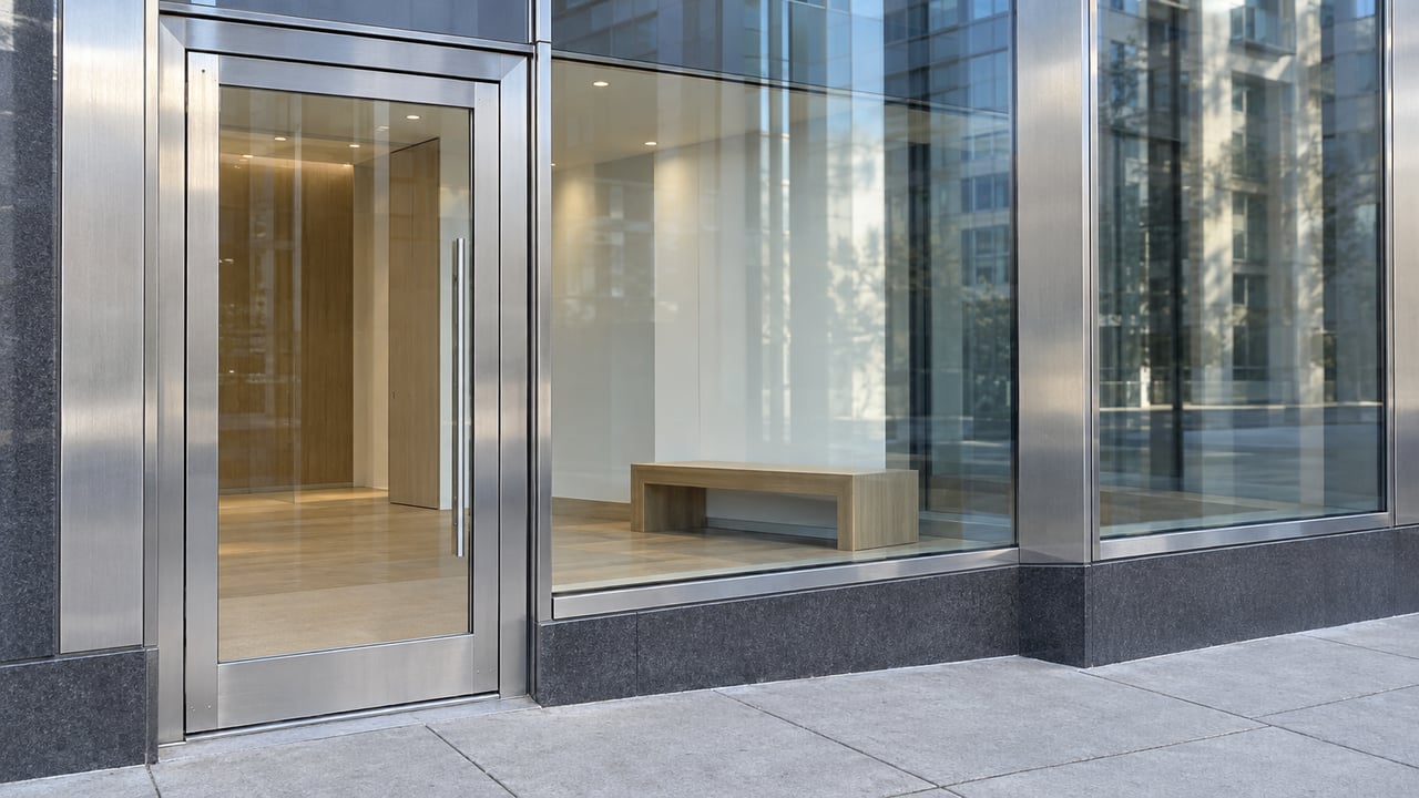 Contemporary condominium entrance with stainless-steel framing, enclosed glass lobby, and pale concrete pavement.