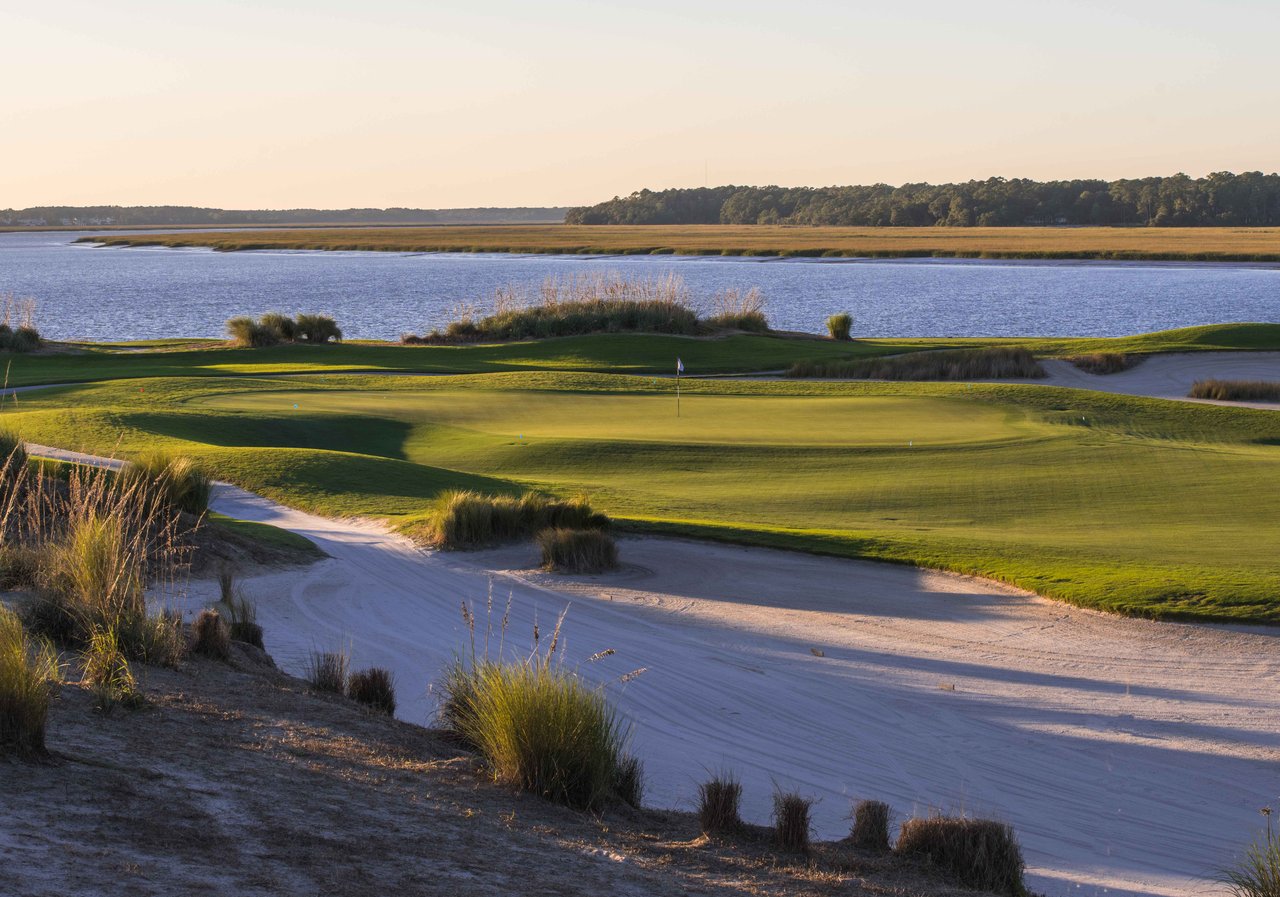 Golf course green and sandy path beside a river and marsh at golden hour.