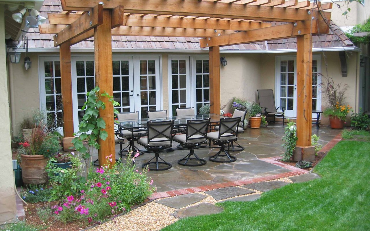 Stone patio with a wooden pergola, outdoor dining set, and colorful flower beds.