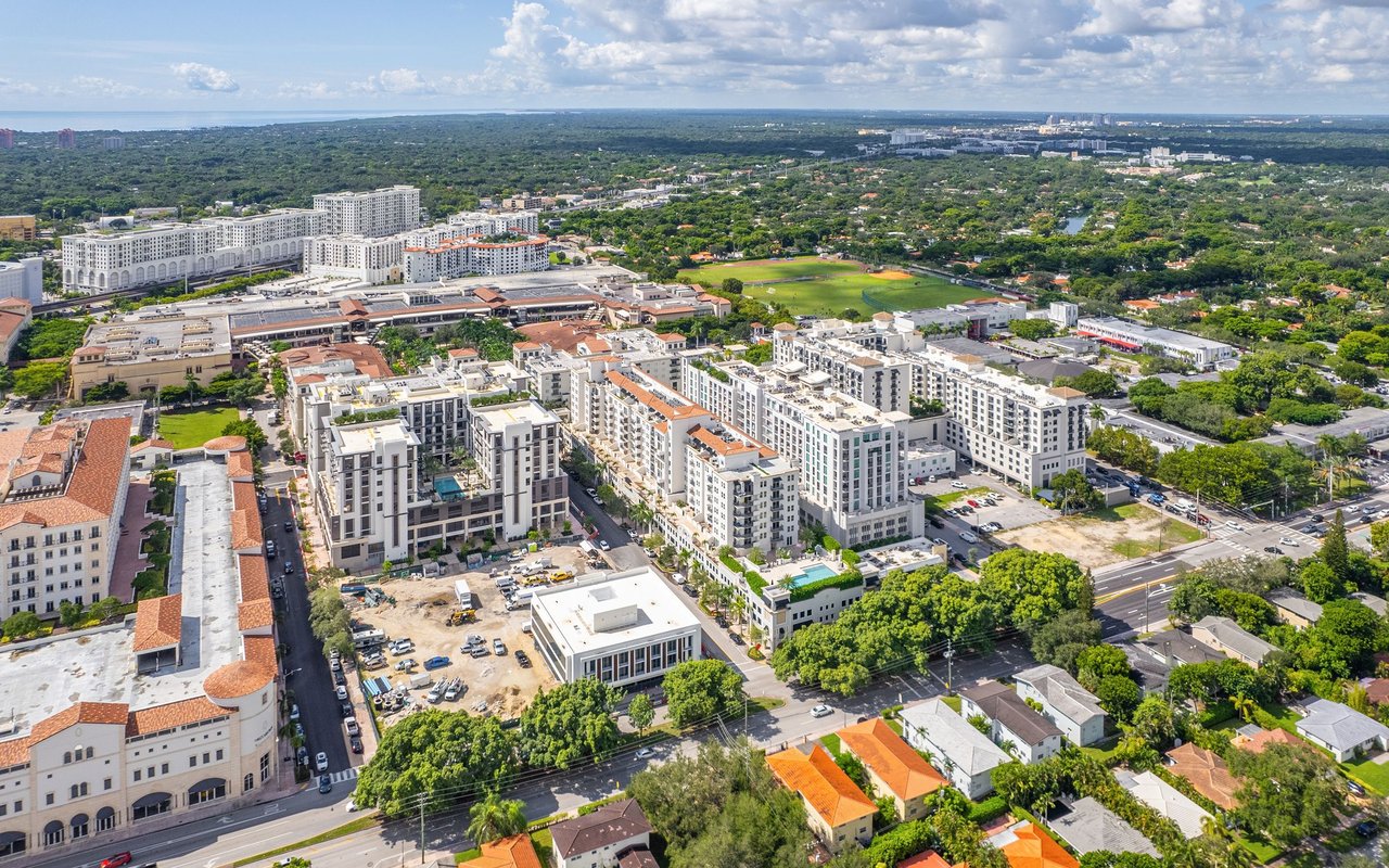 Aerial view of a South Florida district with white mid-rise buildings, tree-lined streets, and a green sports field.