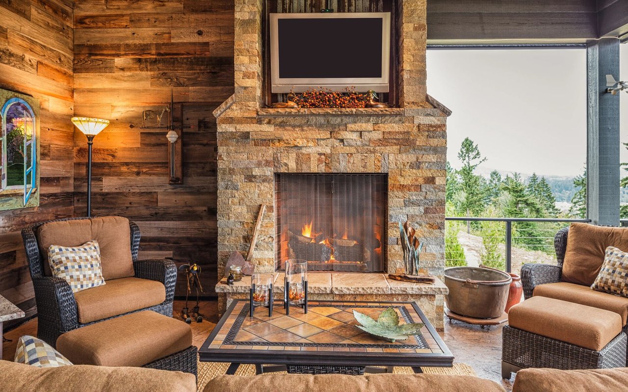 Living room with a stone fireplace, rustic wood walls, and wicker seating beside an outdoor view.