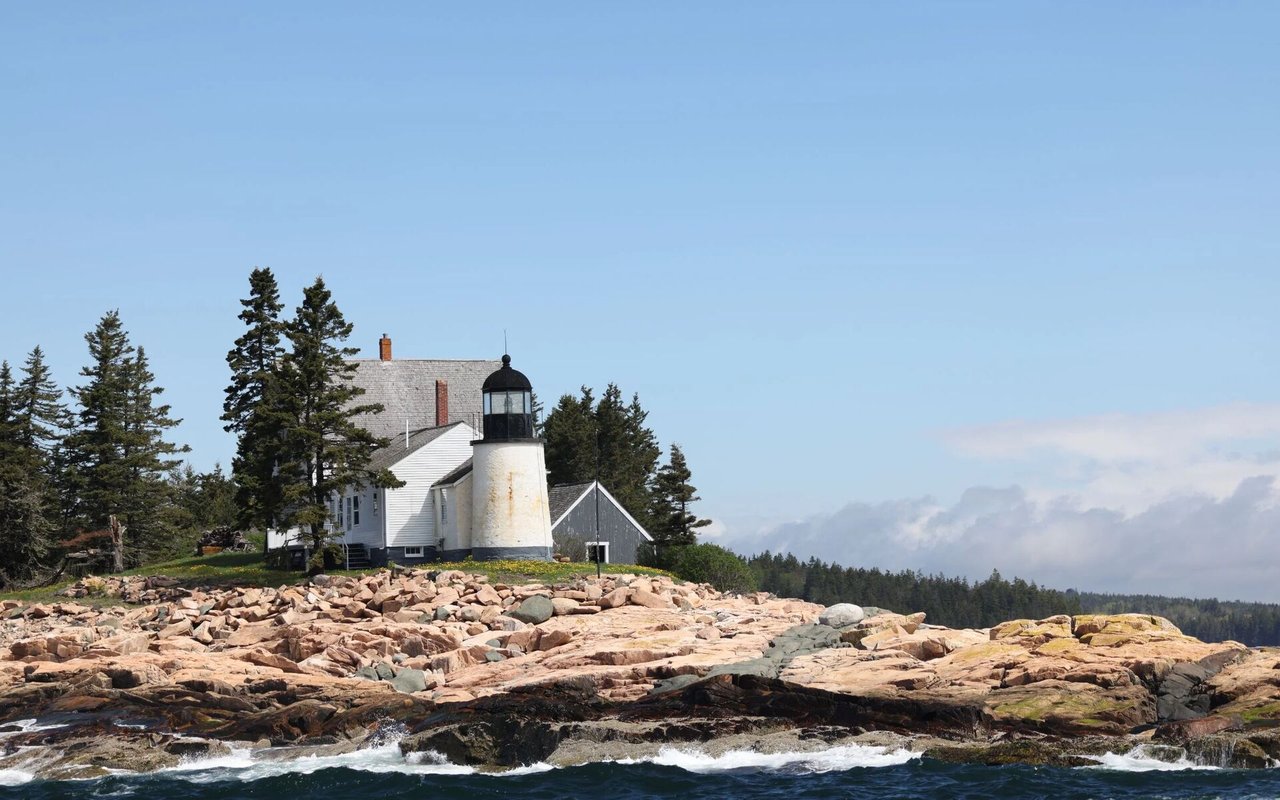 White lighthouse on a rocky coastline, surrounded by evergreens with waves in the foreground.