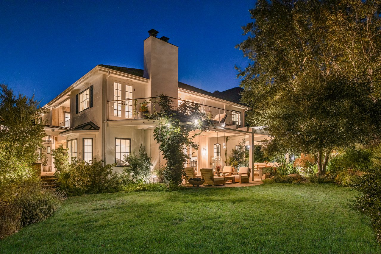 Two-story home at night with a tall chimney and covered patio overlooking a landscaped lawn.