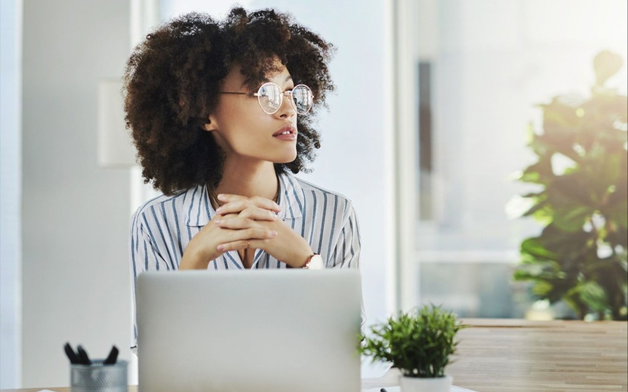 Woman in round glasses looks thoughtfully beside her laptop in a bright office.