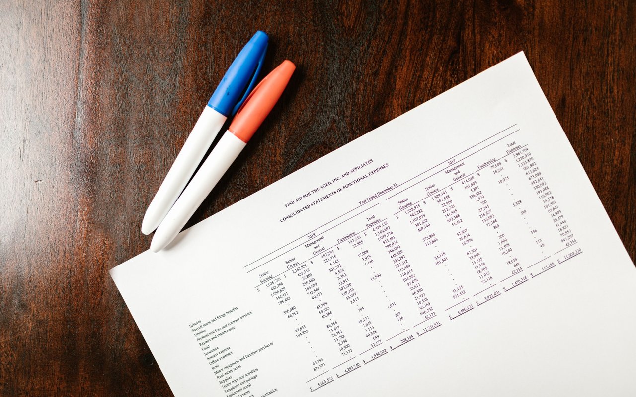 Financial statement with expense tables beside blue and orange markers on a wooden table.