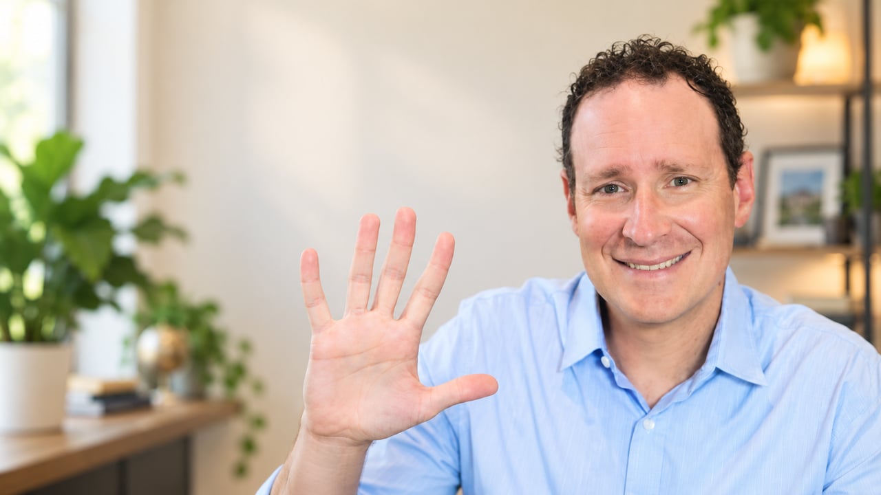 Smiling man in a light blue shirt raises his hand in greeting.