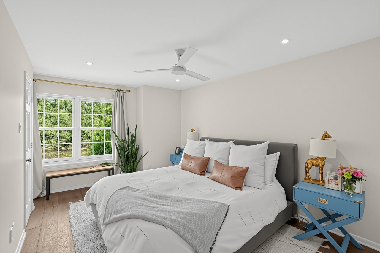 Staged bedroom with a gray upholstered bed, blue bedside tables, and a wide window overlooking greenery.