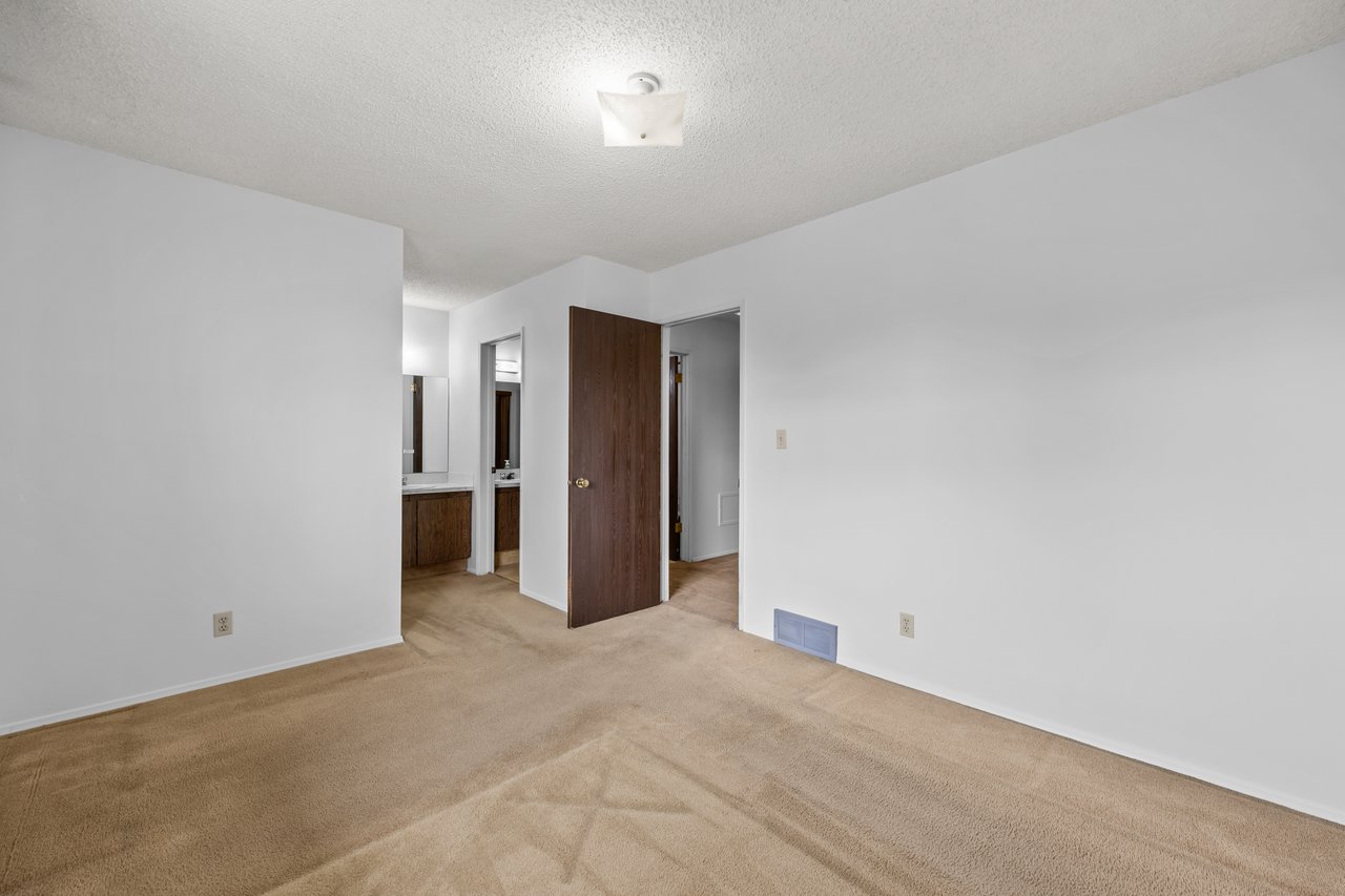 Empty carpeted bedroom with a brown wooden door and bathroom vanity visible through the doorway.