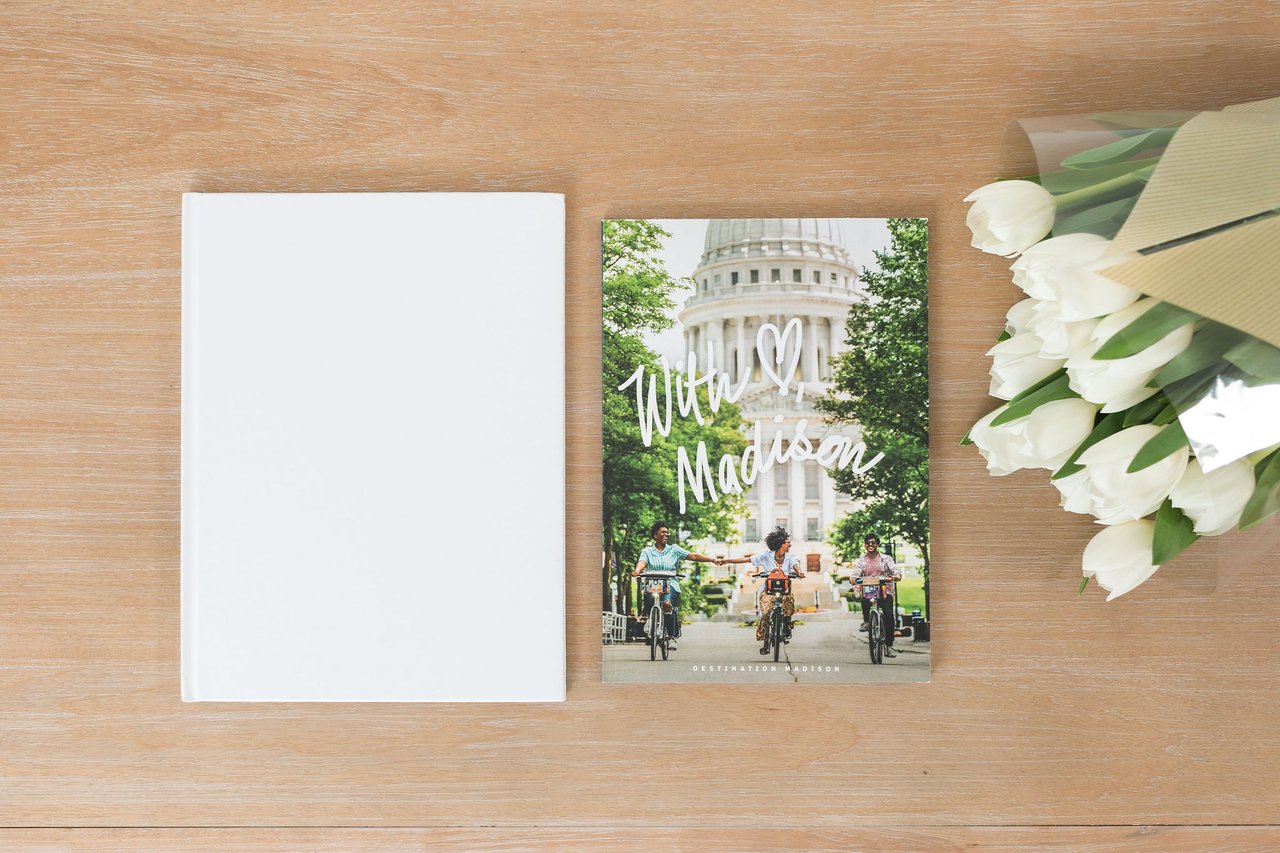 A flat-lay composition showing the Destination Madison magazine featuring the Wisconsin State Capitol and people biking through downtown, alongside a white book and white tulips, representing the kind of research and reference materials that help buyers understand Madison's neighborhoods and geography.