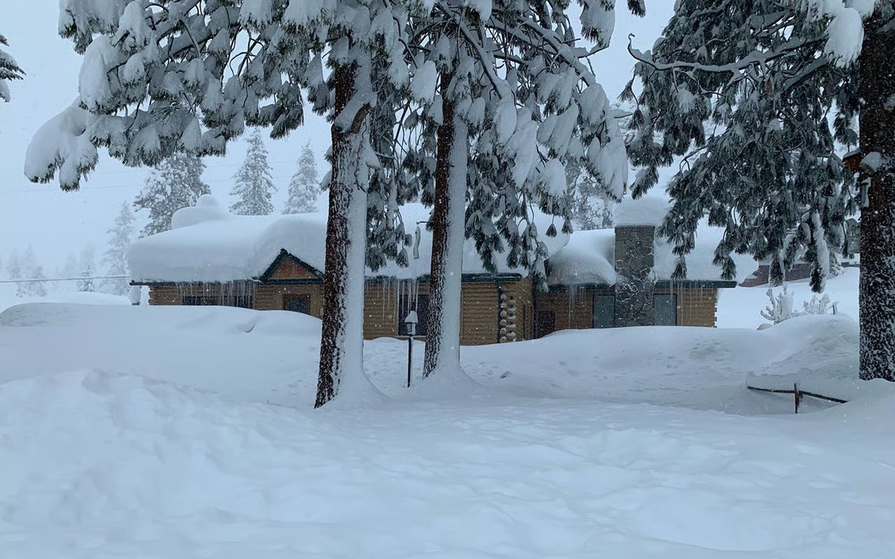 Wooden cabin buried in deep snow, with icicles hanging from its roof beneath snow-covered evergreens.