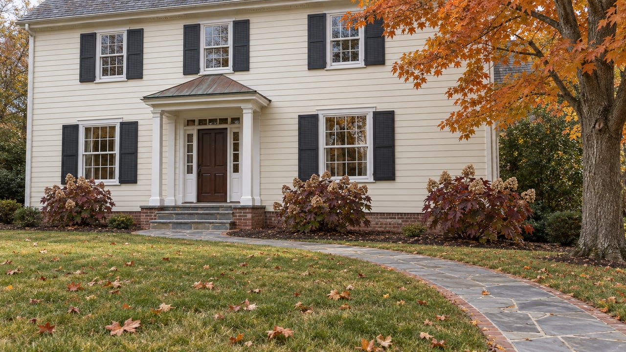 An ivory clapboard home with a copper entry canopy, bluestone walk, and autumn maple leaves scattered across a broad lawn.