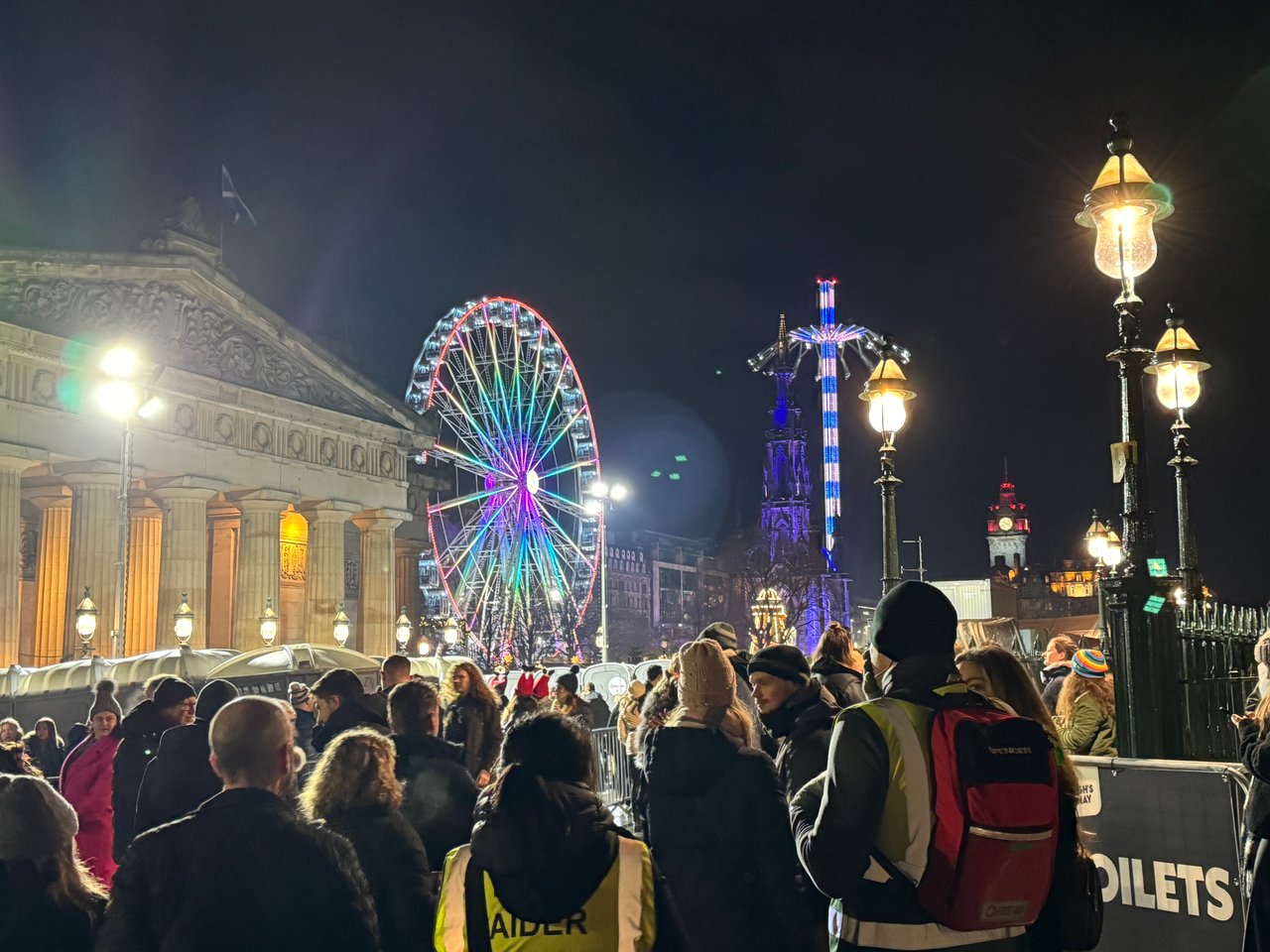 Crowded nighttime city square with a colorful Ferris wheel and illuminated historic buildings.