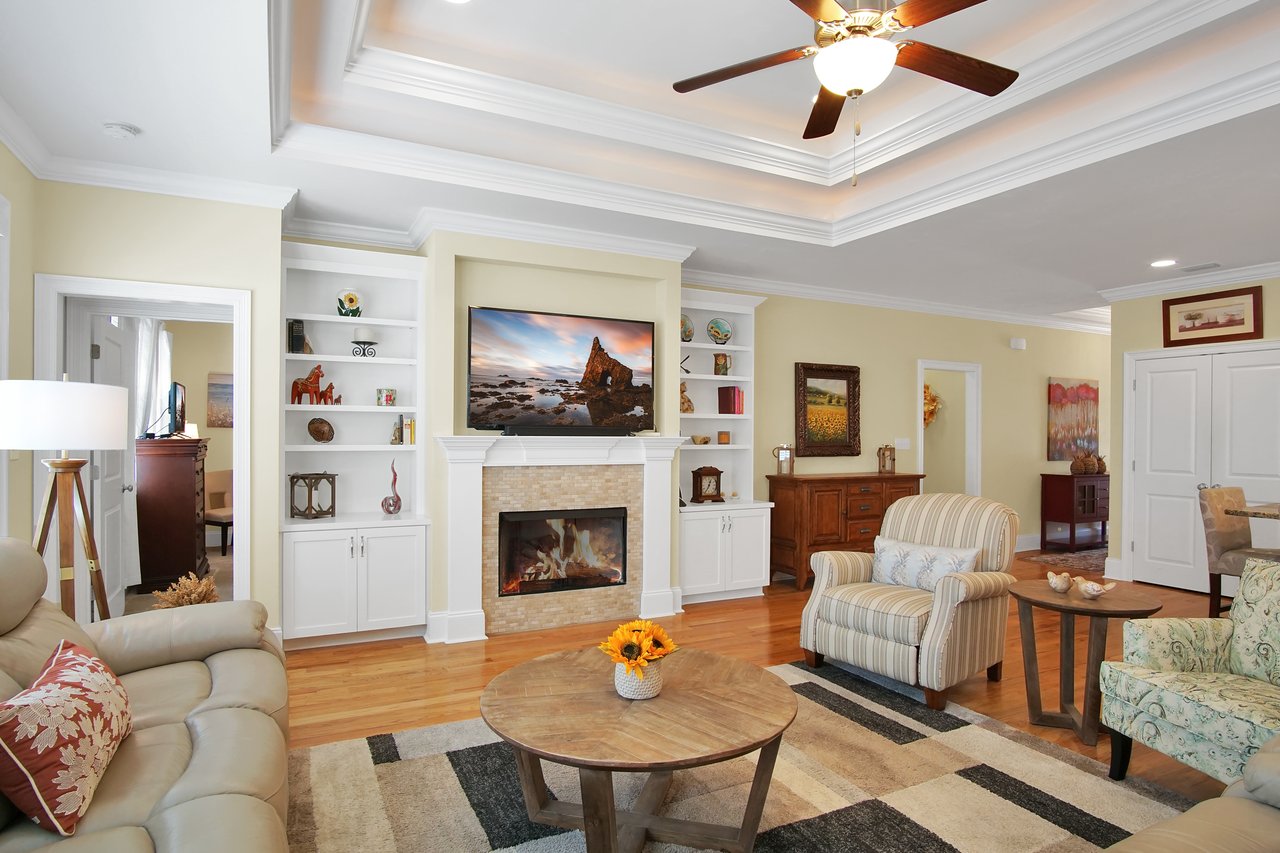 Cozy living room with a fireplace and seaside painting. Beige sofa, striped armchair, and wood coffee table with sunflowers create a warm ambiance.